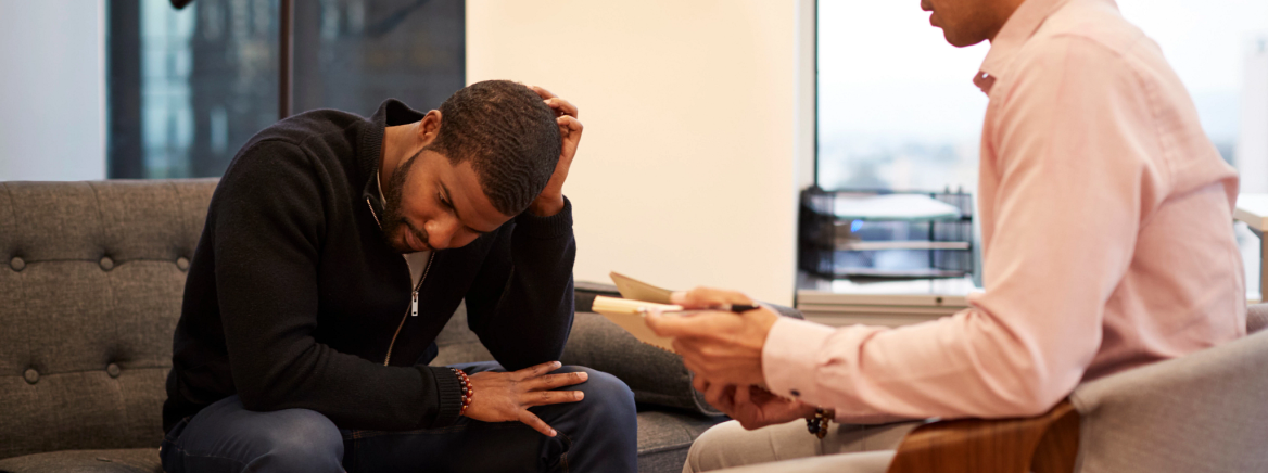 Unhappy Man Sitting On Couch Meeting With Male Counsellor In Office