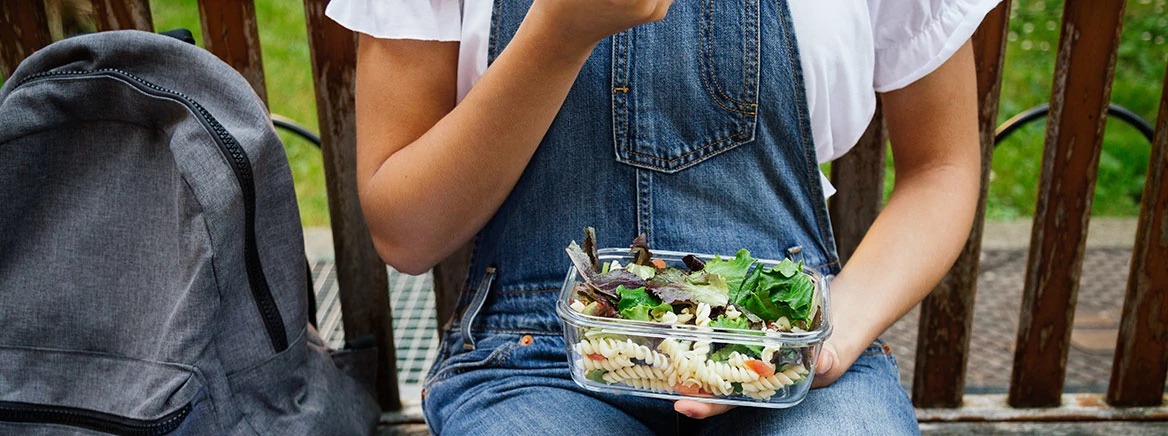 Midsection portrait of a student girl sitting in a bench while eating healthy salad with pasta in a glass lunch box