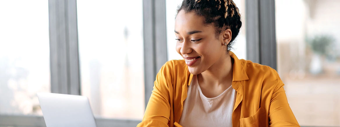 Online education, e-learning. Happy african american young woman in stylish casual clothes, studying remotely, using a laptop, listening to online lecture, taking notes while sitting at home, smiles