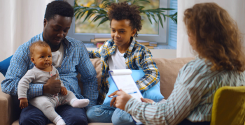 Social worker consulting smiling young single father with kids at home. Case worker woman visiting african man with schoolboy son and baby daughter providing consultation