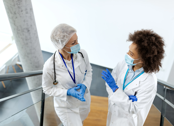 Two female doctors with protective face masks talking while standing in a hallway at medical clinic.