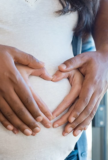 Pregnant woman and her african husband holding hand in heart shape on baby bump. Close up of multiethnic couple making heart shape on the tummy. Loving future couple expecting a baby.