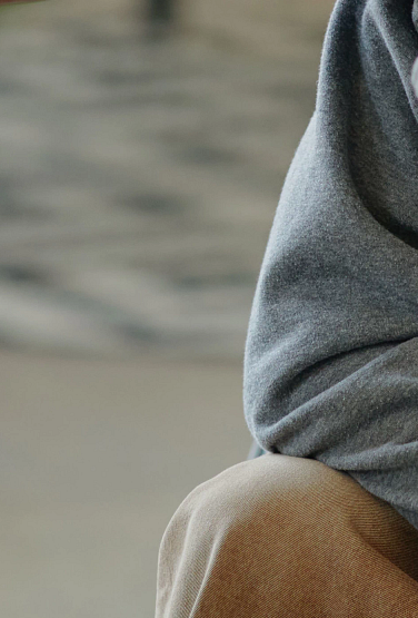 Person comforting distressed teenager sitting with head covered by hood, hand on forehead, supportive gesture in indoor setting, emotional support and empathy visible